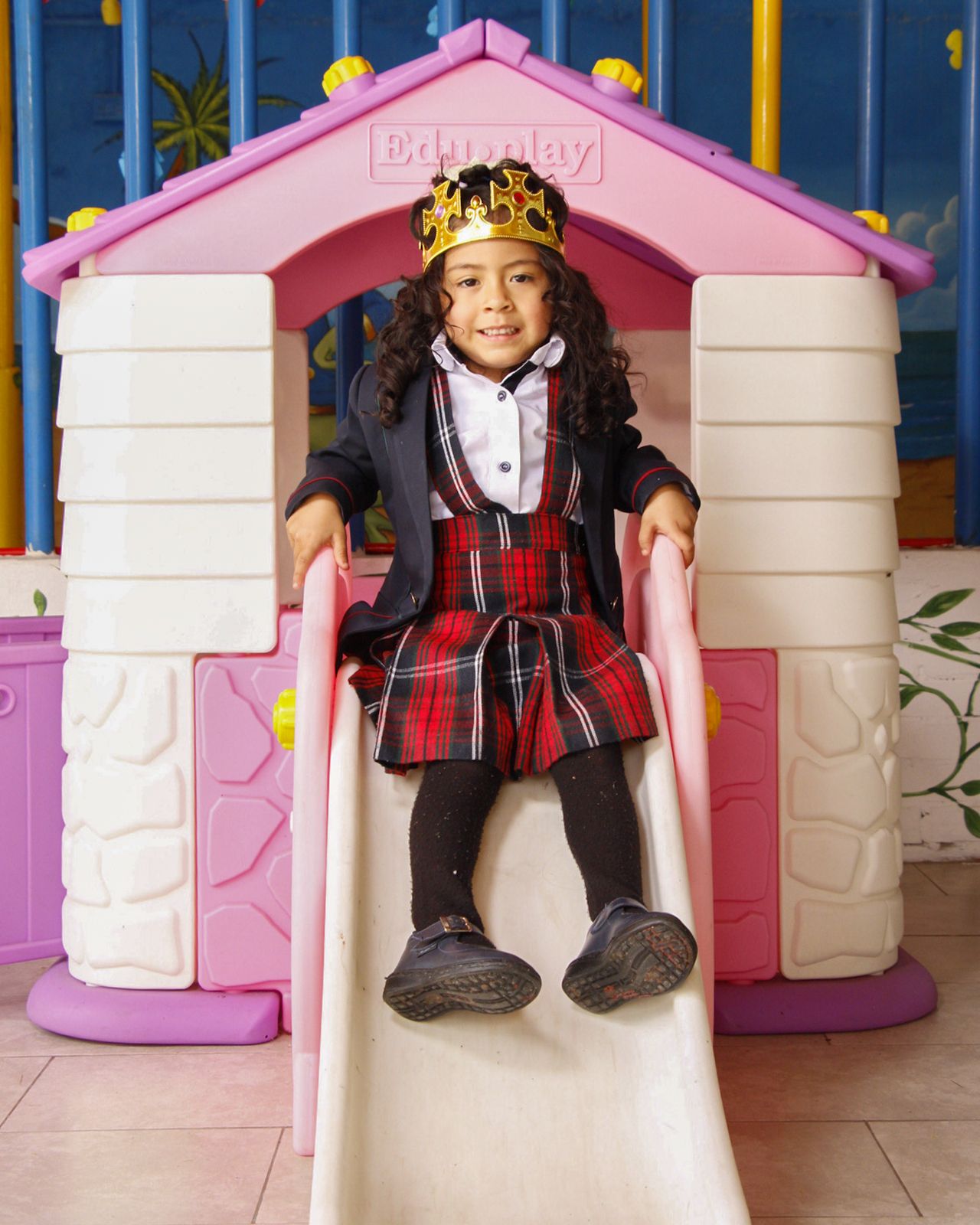 Niña de preescolar con corona sonriendo en el tobogán de la ludoteca